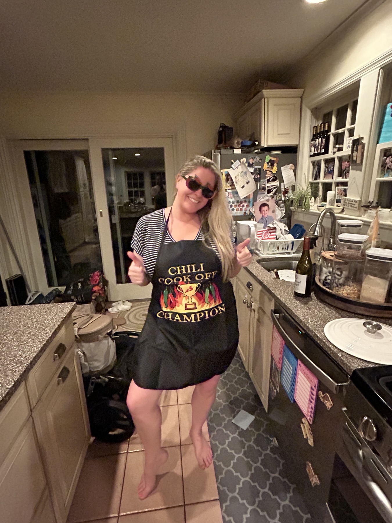 Kathleen in the kitchen at night, wearing sunglasses and a Chili Cook Off Champion apron, giving two thumbs up.