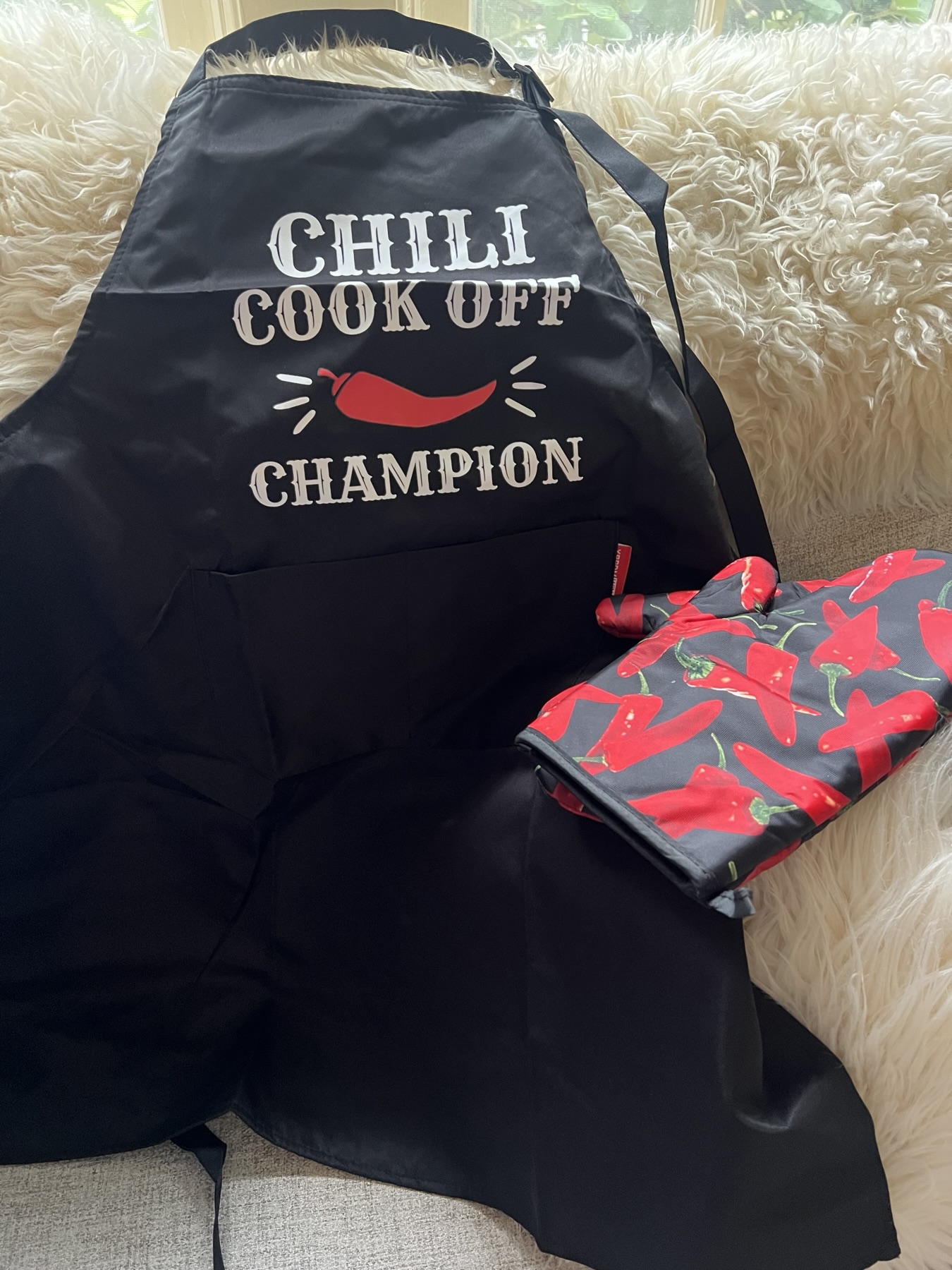 A black Chili Cook Off Champion apron and a red-pepper oven mitt laid out on a cream chair.