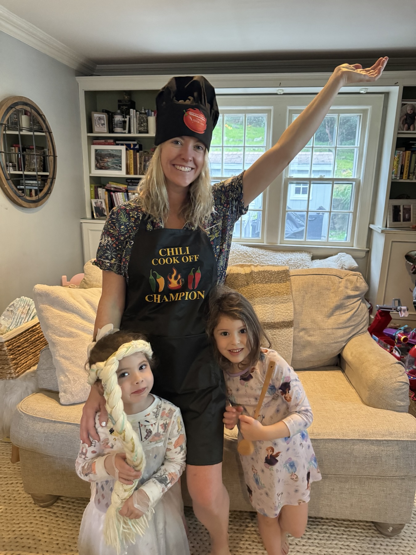 Kathleen in a chef’s hat and Chili Cook Off Champion apron, one arm raised, celebrating with two girls in the living room.